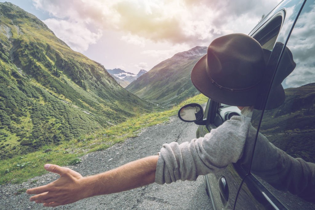 Young man in car on mountain road looks out from window car, outstretched arms for freedom.