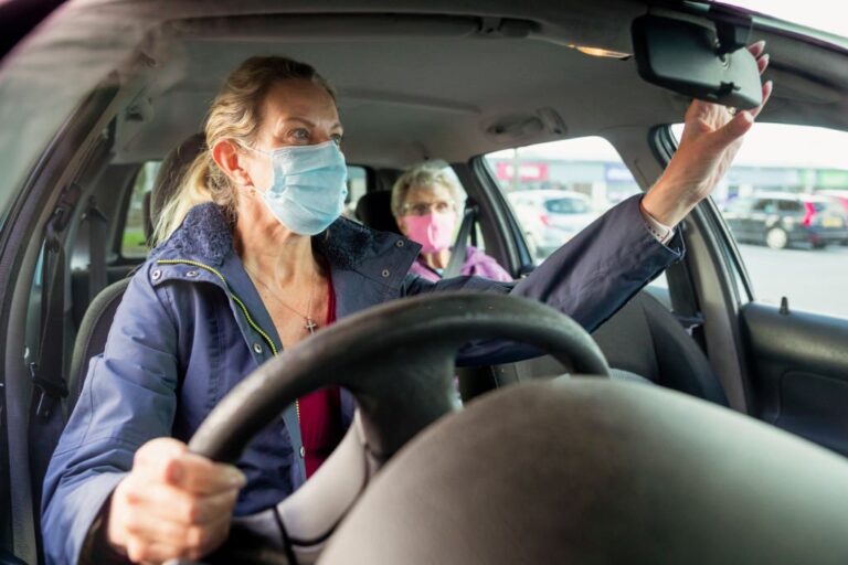 A close-up shot of a mature, Caucasian woman caring for her senior mother. The daughter is adjusting her rear view mirror and they are both wearing face masks to protect themselves and others from Covid-19.