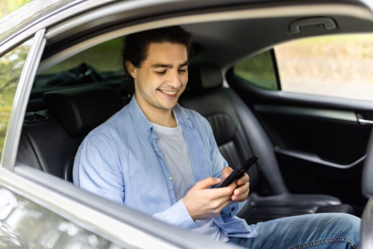Happy smiling business man typing message on phone while sitting in a taxi. Young businessman in formal clothing using smartphone while sitting on back seat in car. Cheerful guy messaging with cell.